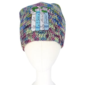 Pode incluir: Um gorro de malha multicolorido com um design floral e de livros. O gorro apresenta um design bordado de três livros com o texto "Just One More Chapter" e um arranjo floral no topo. O chapéu é exibido em uma cabeça de manequim.