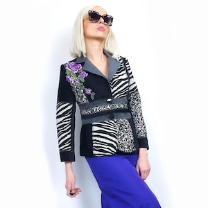 Könnte beinhalten: Ein gemusterter Blazer in Schwarz, Weiß und Grau mit einem violetten Blumenstickerei-Detail. Der Blazer hat einen einzigen Knopfverschluss und einen schwarzen Kragen. Das Model trägt einen blauen Rock.