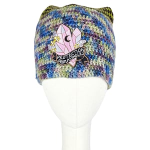 Pode incluir: Um gorro de crochê multicolorido feito à mão com orelhas de gato. O gorro apresenta um patch com um design de cristal rosa, uma lua crescente e o texto "PROTECTION FROM ASSHOLES" em uma faixa preta. O gorro está em uma cabeça de manequim branca.