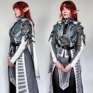 May include: A woman in a grey and white costume with a silver breastplate and matching gauntlets. She has red hair and pointed ears.