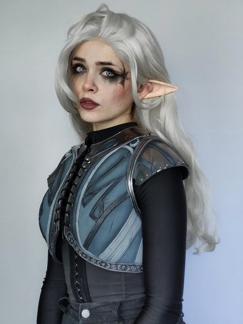 May include: A woman with long, silver hair and pointed ears wears a blue and silver armored vest with a black shirt underneath.