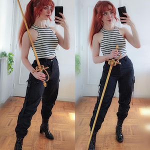 K&ouml;nnte beinhalten: Ein dekoratives Schwert mit goldenem Knauf und langer Holzklinge. Die Person tr&auml;gt ein schwarz-wei&szlig; gestreiftes Oberteil, eine schwarze Hose und schwarze Stiefel. Das Schwert ist das zentrale Objekt im Bild.
