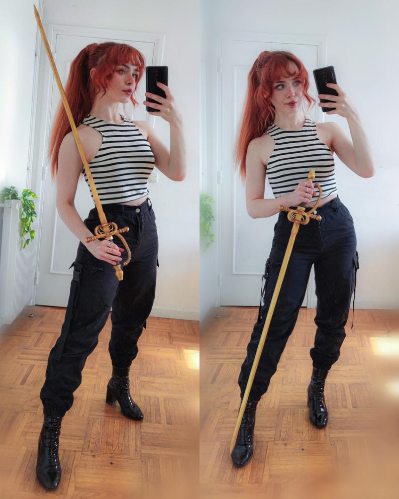 K&ouml;nnte beinhalten: Ein dekoratives Schwert mit goldenem Knauf und langer Holzklinge. Die Person tr&auml;gt ein schwarz-wei&szlig; gestreiftes Oberteil, eine schwarze Hose und schwarze Stiefel. Das Schwert ist das zentrale Objekt im Bild.