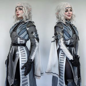 May include: Two figures in fantasy-inspired costumes, both wearing white and grey armor with intricate details. They have long white hair and pointed ears. The figures are posed in a symmetrical fashion, with one facing left and the other facing right.