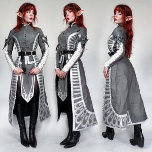 May include: A gray and silver costume with intricate details and a high collar. The costume features a long skirt with a silver trim and a belt with a silver buckle. The costume is worn by a person with red hair and elf ears.