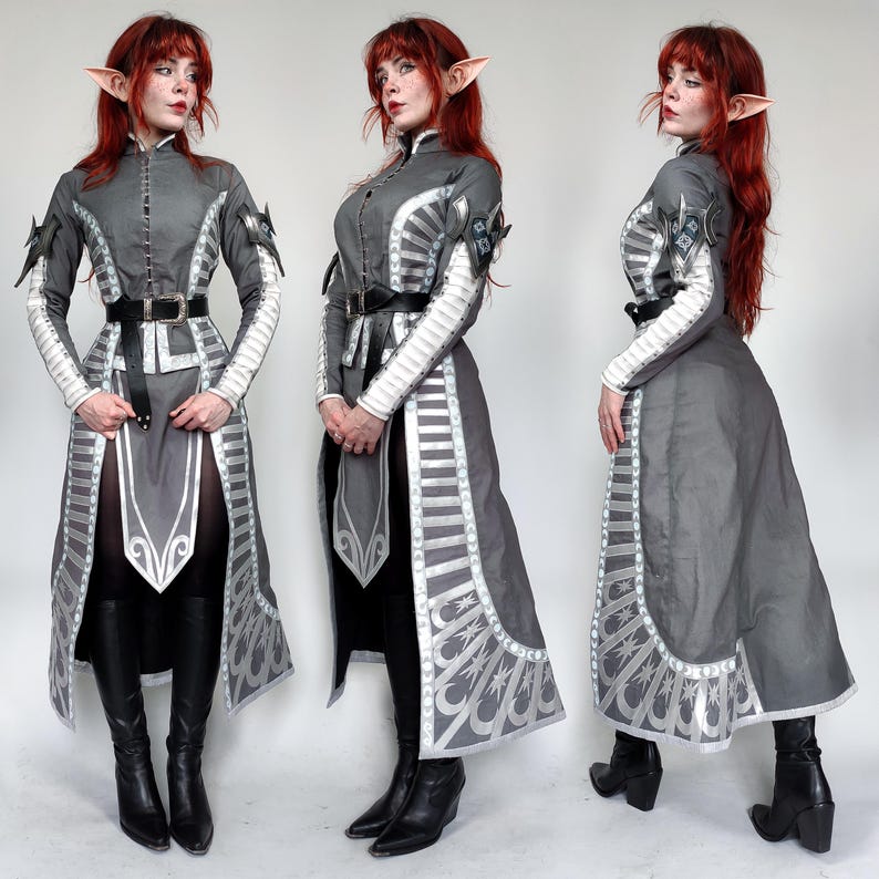 May include: A gray and silver costume with intricate details and a high collar. The costume features a long skirt with a silver trim and a belt with a silver buckle. The costume is worn by a person with red hair and elf ears.