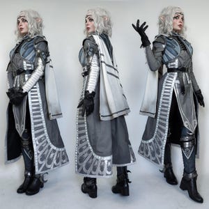 May include: A woman in a grey and white costume with silver accents. She is wearing a long, flowing cape and a belt with a silver buckle. She has long, white hair and is wearing a silver crown. The costume is detailed with intricate designs and patterns.