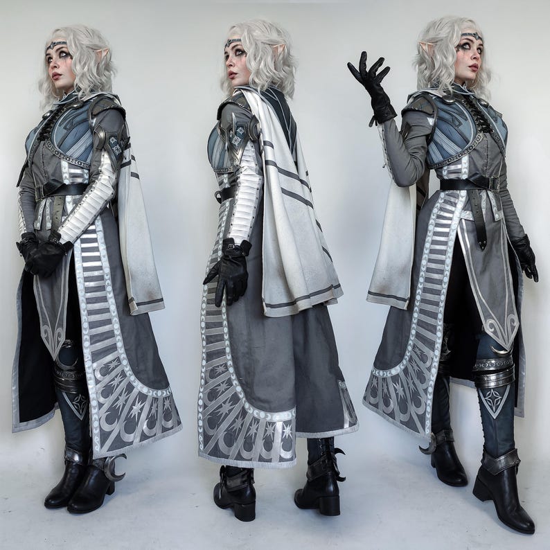 May include: A woman in a grey and white costume with silver accents. She is wearing a long, flowing cape and a belt with a silver buckle. She has long, white hair and is wearing a silver crown. The costume is detailed with intricate designs and patterns.