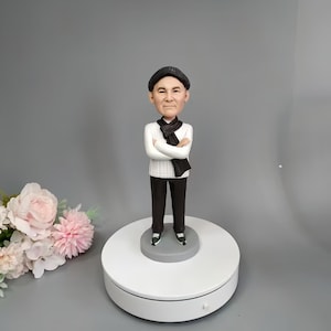 Könnte beinhalten: Eine personalisierte Bobblehead-Figur einer Person, die einen weißen Pullover, eine schwarze Hose, einen schwarzen Schal und einen schwarzen Hut trägt. Die Figur steht auf einem weißen runden Sockel.