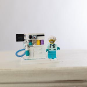 May include: A Lego model of a medical machine with gauges, dials, and a screen, next to a Lego figure of a doctor wearing a surgical cap and scrubs. The machine has white, teal, and black components.