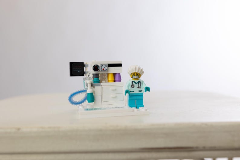 May include: A Lego model of a medical machine with gauges, dials, and a screen, next to a Lego figure of a doctor wearing a surgical cap and scrubs. The machine has white, teal, and black components.