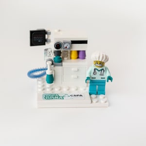 May include: A Lego-style medical scene featuring a white and teal medical machine with gauges, tubes, and colorful vials. A Lego figure in teal scrubs and a white cap stands nearby. The base of the machine has the text "CSNA CSPA".