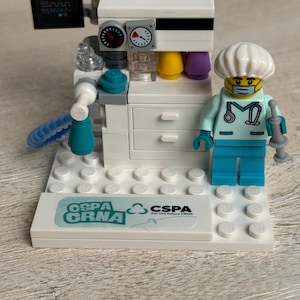 May include: A white Lego medical machine with a monitor, gauges, and colorful tubes. A Lego figure in a medical uniform and mask stands beside the machine. The base has the text "GSPA CRNA" and "CSPA For The Future CRNA".