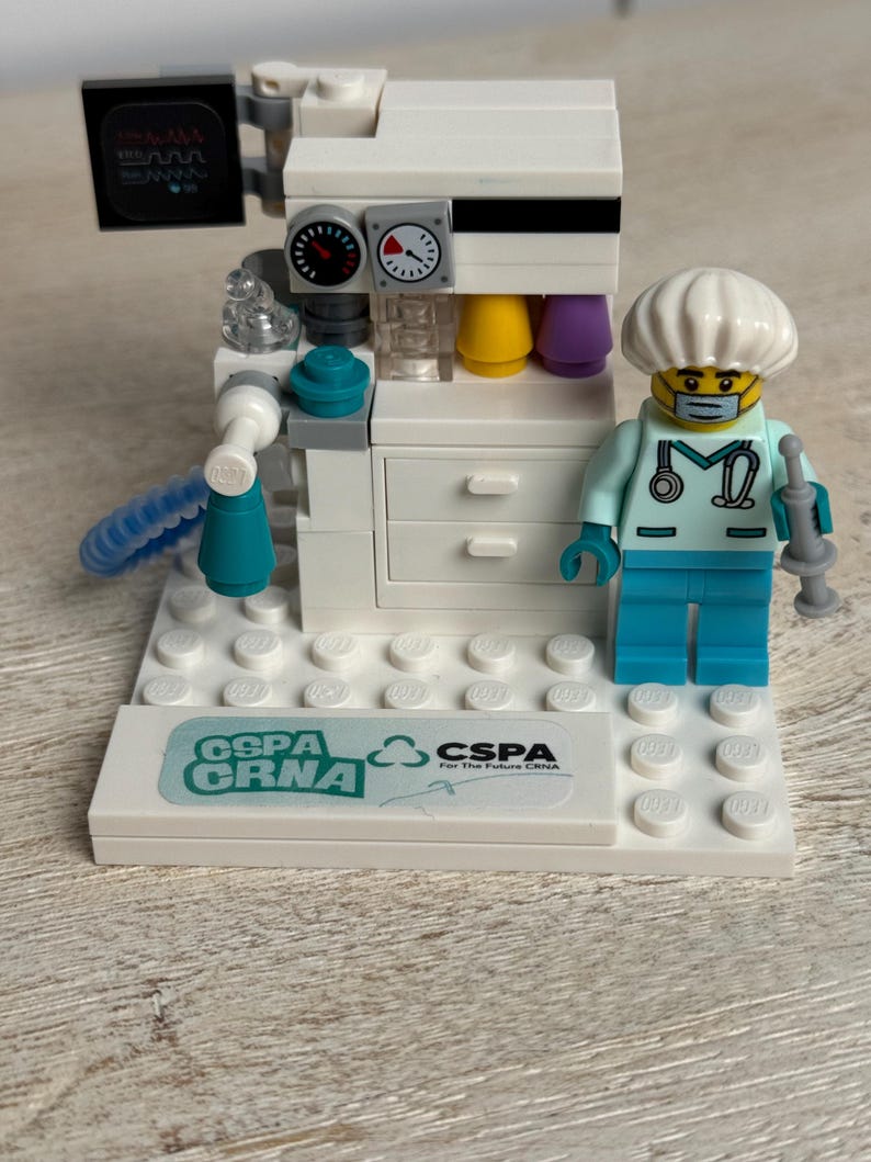 May include: A white Lego medical machine with a monitor, gauges, and colorful tubes. A Lego figure in a medical uniform and mask stands beside the machine. The base has the text "GSPA CRNA" and "CSPA For The Future CRNA".