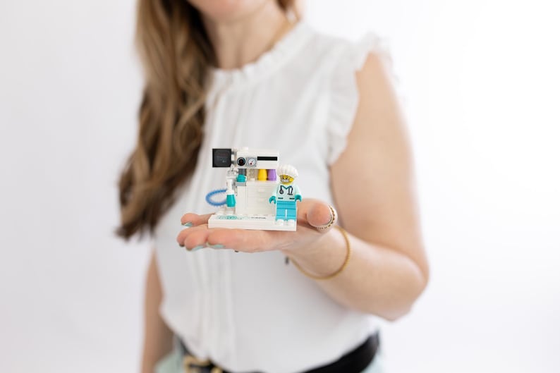 May include: A Lego model of an operating room with a miniature medical professional. The model features a white base with a teal and blue medical machine, and a doctor figurine in blue scrubs and a white cap. The model is held in a person's hand.