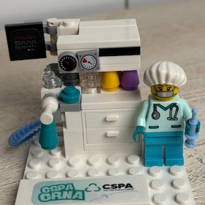 May include: A Lego model of a medical machine with a doctor figurine. The machine is white with black and yellow details, and the doctor is wearing a light blue uniform and a white cap. The base has the text "CSPA CRNA".