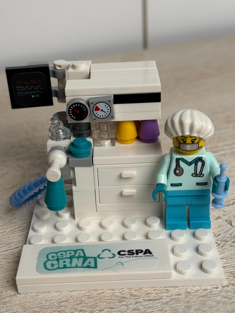 May include: A Lego model of a medical machine with a doctor figurine. The machine is white with black and yellow details, and the doctor is wearing a light blue uniform and a white cap. The base has the text "CSPA CRNA".
