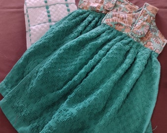 May include: A green tea towel dress with a floral print fabric sewn into the bodice. The dress is made from two tea towels and has a white and green checkered tea towel on the side.