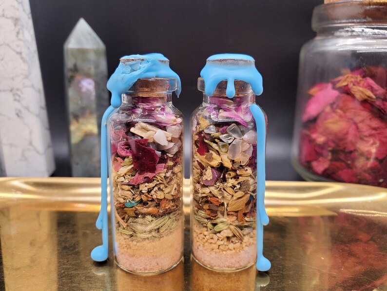 K&ouml;nnte beinhalten: Zwei kleine Glasflaschen gef&uuml;llt mit Schichten aus getrockneten Kr&auml;utern, Kristallen und Sand. Die Flaschen sind mit Korken und blauem Wachs verschlossen. Die Flaschen stehen auf einem goldenen Tablett, mit einem Hintergrund aus Kristallen und einem Glas getrockneter Blumen.