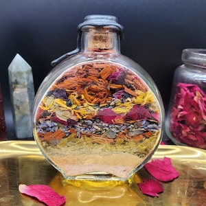 Könnte beinhalten: Eine runde Glasflasche gefüllt mit Schichten aus getrockneten Kräutern, Blumen und Holzspänen. Die Flasche hat einen Korkverschluss und steht auf einem goldenen Tablett.