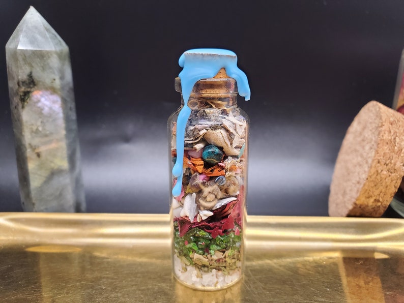 K&ouml;nnte beinhalten: Eine kleine Glasflasche gef&uuml;llt mit Schichten aus getrockneten Kr&auml;utern, Blumen und Kristallen. Die Flasche ist mit einem Korken verschlossen und mit blauem Wachs bedeckt. Der Inhalt ist durch das klare Glas sichtbar.