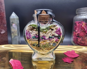 Puede incluir: Una botella de vidrio en forma de corazón llena de hierbas secas, flores y cristales. La botella tiene un tapón de corcho sellado con cera negra. El contenido incluye elementos verdes, morados y beige. La botella está sobre una bandeja dorada.