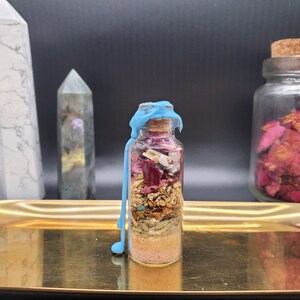 K&ouml;nnte beinhalten: Eine kleine Glasflasche gef&uuml;llt mit Schichten aus getrockneten Kr&auml;utern, Bl&uuml;tenbl&auml;ttern und Kristallen, verschlossen mit einem Korken und blauem Wachs. Die Flasche steht auf einem goldenen Tablett, mit Kristallen und einem Glas Rosenbl&uuml;ten im Hintergrund.