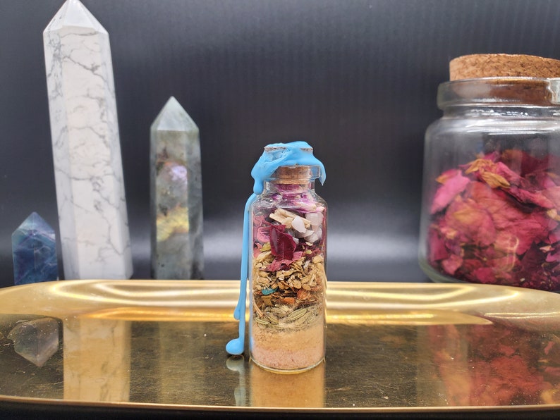 K&ouml;nnte beinhalten: Eine kleine Glasflasche gef&uuml;llt mit Schichten aus getrockneten Kr&auml;utern, Bl&uuml;tenbl&auml;ttern und Kristallen, verschlossen mit einem Korken und blauem Wachs. Die Flasche steht auf einem goldenen Tablett, mit Kristallen und einem Glas Rosenbl&uuml;ten im Hintergrund.