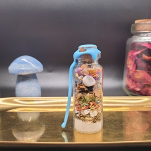 K&ouml;nnte beinhalten: Eine kleine Glasflasche gef&uuml;llt mit bunten getrockneten Kr&auml;utern, Kristallen und anderen Zutaten, verschlossen mit einem Korken und blauem Wachs. Die Flasche steht auf einem goldenen Tablett, mit einem blauen Pilz und anderen dekorativen Gegenst&auml;nden im Hintergrund.