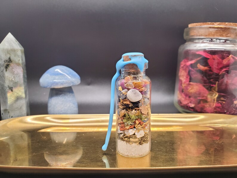 K&ouml;nnte beinhalten: Eine kleine Glasflasche gef&uuml;llt mit bunten getrockneten Kr&auml;utern, Kristallen und anderen Zutaten, verschlossen mit einem Korken und blauem Wachs. Die Flasche steht auf einem goldenen Tablett, mit einem blauen Pilz und anderen dekorativen Gegenst&auml;nden im Hintergrund.