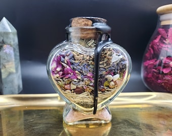 Pode incluir: Um frasco de vidro em forma de coração cheio de ervas secas, flores e cristais. O frasco tem uma rolha de cortiça e é selado com cera preta. O frasco está sobre uma bandeja dourada.