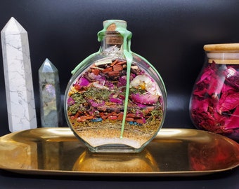 May include: A round glass bottle filled with layers of dried herbs, flowers, and crystals. The bottle is sealed with a cork and has a green wax seal. The bottle is sitting on a gold tray.