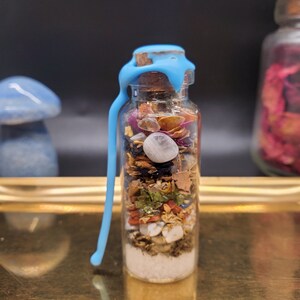 K&ouml;nnte beinhalten: Kleine Glasflasche gef&uuml;llt mit Schichten aus getrockneten Kr&auml;utern, Kristallen und anderen Zutaten. Die Flasche ist mit einem Korken und blauem Wachs verschlossen. Im Hintergrund befindet sich ein blaues pilzf&ouml;rmiges Objekt.