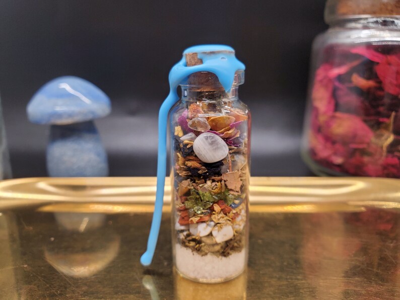 K&ouml;nnte beinhalten: Kleine Glasflasche gef&uuml;llt mit Schichten aus getrockneten Kr&auml;utern, Kristallen und anderen Zutaten. Die Flasche ist mit einem Korken und blauem Wachs verschlossen. Im Hintergrund befindet sich ein blaues pilzf&ouml;rmiges Objekt.