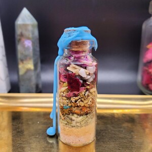 K&ouml;nnte beinhalten: Eine kleine Glasflasche gef&uuml;llt mit Schichten aus getrockneten Kr&auml;utern, Kristallen und Blumen, verschlossen mit einem Korken und blauem Wachs. Die Flasche steht auf einem goldenen Tablett, mit anderen dekorativen Gegenst&auml;nden im Hintergrund.