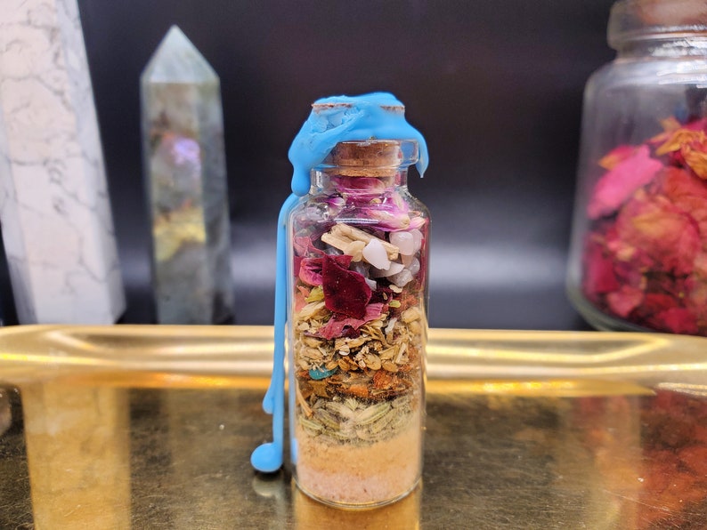 K&ouml;nnte beinhalten: Eine kleine Glasflasche gef&uuml;llt mit Schichten aus getrockneten Kr&auml;utern, Kristallen und Blumen, verschlossen mit einem Korken und blauem Wachs. Die Flasche steht auf einem goldenen Tablett, mit anderen dekorativen Gegenst&auml;nden im Hintergrund.