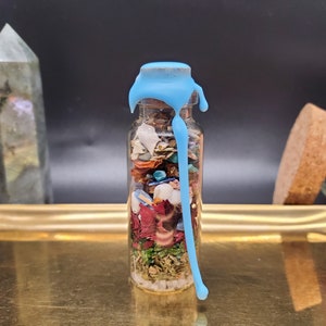 K&ouml;nnte beinhalten: Eine kleine Glasflasche gef&uuml;llt mit bunten getrockneten Kr&auml;utern, Bl&uuml;tenbl&auml;ttern und Kristallen, versiegelt mit hellblauem Wachs. Die Flasche steht auf einem goldenen Tablett, mit einem Kristall und einem Glas Rosenbl&uuml;ten im Hintergrund.
