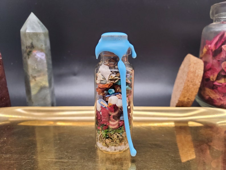 K&ouml;nnte beinhalten: Eine kleine Glasflasche gef&uuml;llt mit bunten getrockneten Kr&auml;utern, Bl&uuml;tenbl&auml;ttern und Kristallen, versiegelt mit hellblauem Wachs. Die Flasche steht auf einem goldenen Tablett, mit einem Kristall und einem Glas Rosenbl&uuml;ten im Hintergrund.