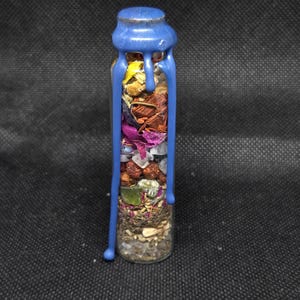 K&ouml;nnte beinhalten: Eine kleine Glasflasche gef&uuml;llt mit getrockneten Kr&auml;utern, Samen und Kristallen. Die Flasche ist mit blauem Wachs versiegelt, das an den Seiten herunterl&auml;uft. Durch das klare Glas sind die Inhalte mit verschiedenen Farben und Texturen sichtbar.