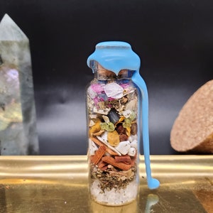 K&ouml;nnte beinhalten: Eine transparente Glasflasche gef&uuml;llt mit getrockneten Kr&auml;utern, Kristallen und Pflanzen, versiegelt mit hellblauem Wachs. Die Flasche steht auf einem goldenen Tablett vor schwarzem Hintergrund. Inhalt: wei&szlig;e, rote, gr&uuml;ne und braune Elemente.