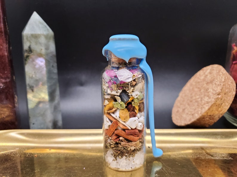 K&ouml;nnte beinhalten: Eine transparente Glasflasche gef&uuml;llt mit getrockneten Kr&auml;utern, Kristallen und Pflanzen, versiegelt mit hellblauem Wachs. Die Flasche steht auf einem goldenen Tablett vor schwarzem Hintergrund. Inhalt: wei&szlig;e, rote, gr&uuml;ne und braune Elemente.