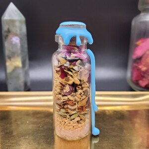 K&ouml;nnte beinhalten: Eine kleine Glasflasche gef&uuml;llt mit getrockneten Kr&auml;utern, Kristallen und rosa Sand. Die Flasche ist mit hellblauem Wachs versiegelt, das an der Seite herunterl&auml;uft. Die Flasche steht auf einem goldenen Tablett, mit anderen dekorativen Gegenst&auml;nden im Hintergrund.