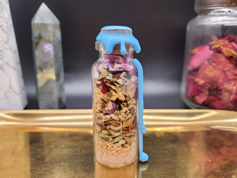 K&ouml;nnte beinhalten: Eine kleine Glasflasche gef&uuml;llt mit getrockneten Kr&auml;utern, Kristallen und rosa Sand. Die Flasche ist mit hellblauem Wachs versiegelt, das an der Seite herunterl&auml;uft. Die Flasche steht auf einem goldenen Tablett, mit anderen dekorativen Gegenst&auml;nden im Hintergrund.