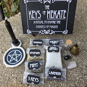 Könnte beinhalten: Eine Sammlung von Ritualgegenständen zur Anrufung der Göttin Hekate. Enthält eine schwarze Kerze, ein Pentakel, kleine Beutel mit Kräutern und Kristallen, eine Glocke und ein Schild mit der Aufschrift "The Keys of Hekate". Die Gegenstände sind auf einer strukturierten Oberfläche angeordnet.