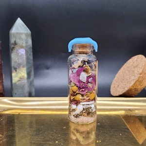 K&ouml;nnte beinhalten: Kleine Glasflasche gef&uuml;llt mit Schichten aus getrockneten Blumen, Kr&auml;utern und Kristallen. Die Flasche hat einen Korken mit blauem Kunststoffverschluss. Inhalt: rosa Rosenbl&uuml;ten, gelbe Kamille und wei&szlig;e Muscheln. Auf einem goldenen Tablett.