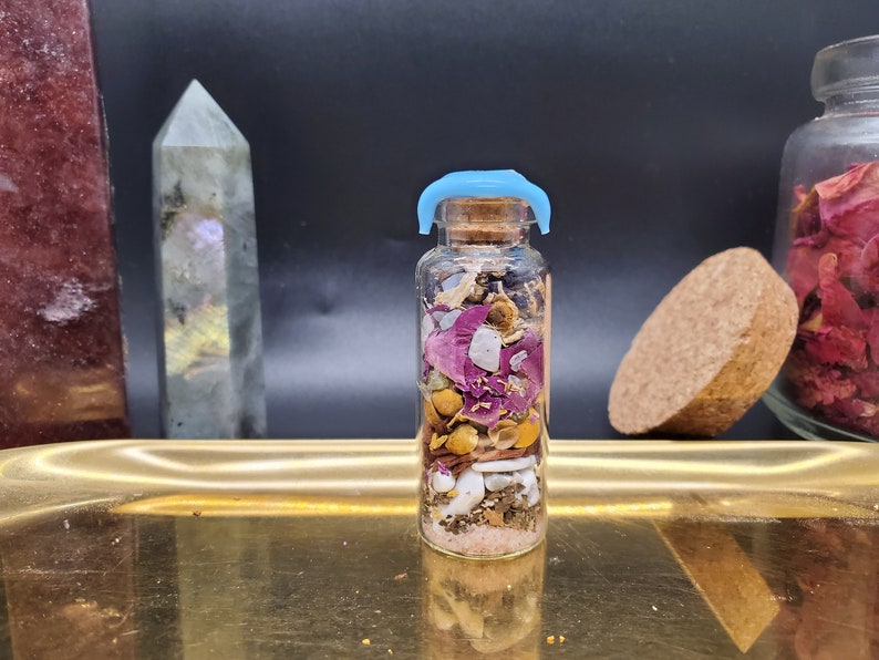 K&ouml;nnte beinhalten: Kleine Glasflasche gef&uuml;llt mit Schichten aus getrockneten Blumen, Kr&auml;utern und Kristallen. Die Flasche hat einen Korken mit blauem Kunststoffverschluss. Inhalt: rosa Rosenbl&uuml;ten, gelbe Kamille und wei&szlig;e Muscheln. Auf einem goldenen Tablett.