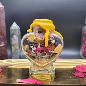 May include: A heart-shaped glass jar filled with dried herbs, flowers, and crystals. The jar has a yellow wax seal on top. The jar is sitting on a gold tray with rose petals.