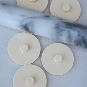 May include: Five off-white round silicone baking mats with a small knob in the center. The mats are arranged in a semi-circle around a marble rolling pin.