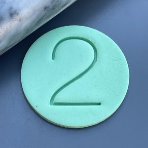 Può includere: Un timbro rotondo in argilla verde chiaro con il numero 2 in rilievo al centro.