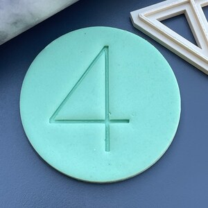 Può includere: Un disco rotondo di argilla verde chiaro con il numero "4" in rilievo sulla superficie.